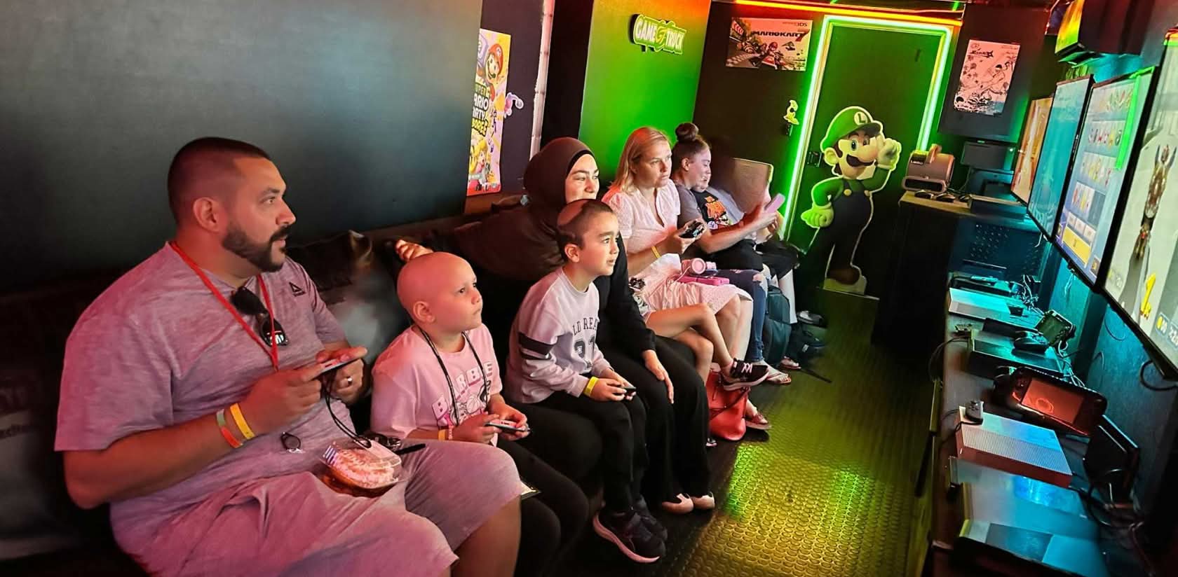 a group of parents and children are seated on bench inside a mobile video game truck and are intently looking at the screens on the opposite wall while holding video game controls