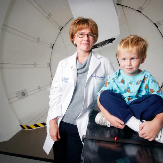 Dr. Nancy Mendenhall with child wearing a patient gown seated cross-legged on the proton treatment table in the gantry treatment room