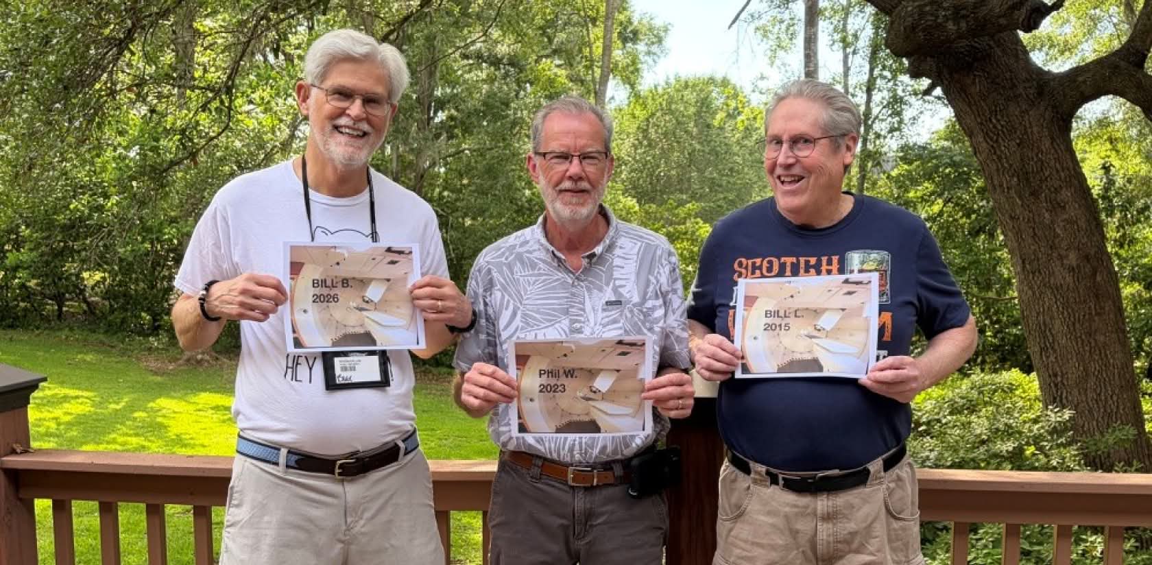 three men smiling and standing together on a wooden deck with a grassy lawn and trees in the background, each holding a photo of a proton treatment room printed with their names and the dates they were treated
