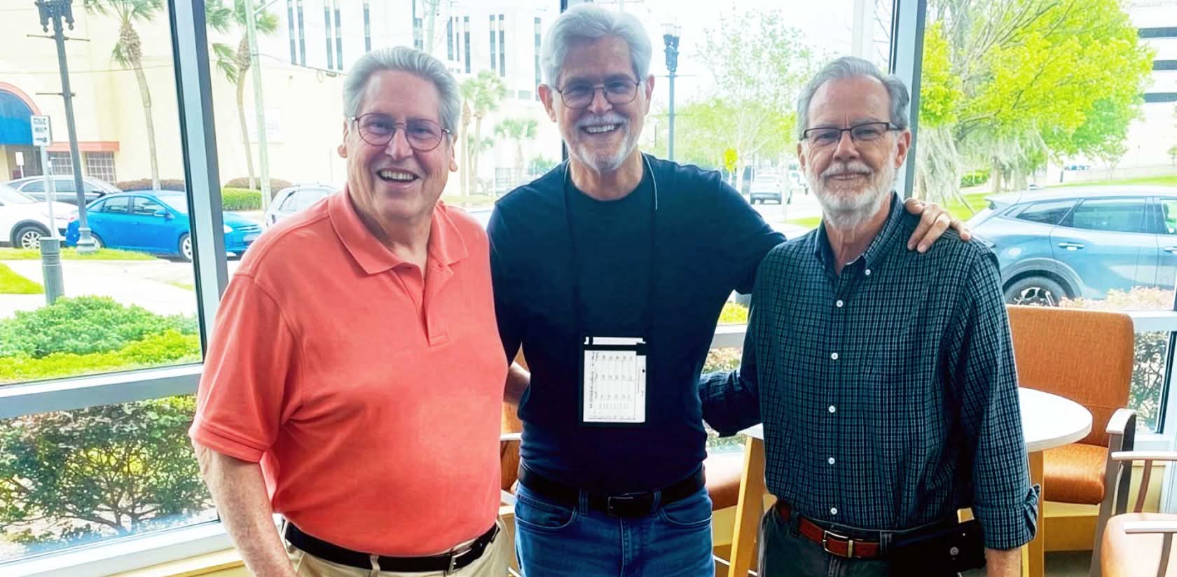 three men smiling and standing together with a large window and street scene behind them
