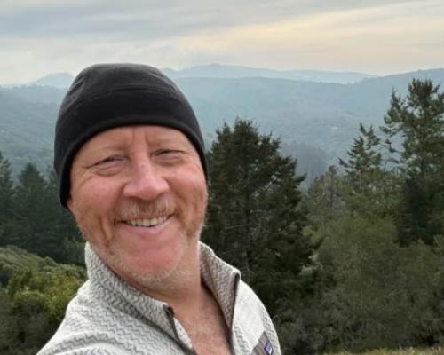 John Ogle takes a selfie while enjoying an active lifestyle after cancer treatment. Hills and trees are in the background and mountains are in the distance 